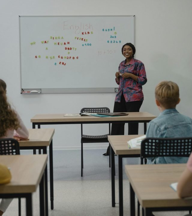 A diverse group of students in an English class with a smiling teacher at the whiteboard.